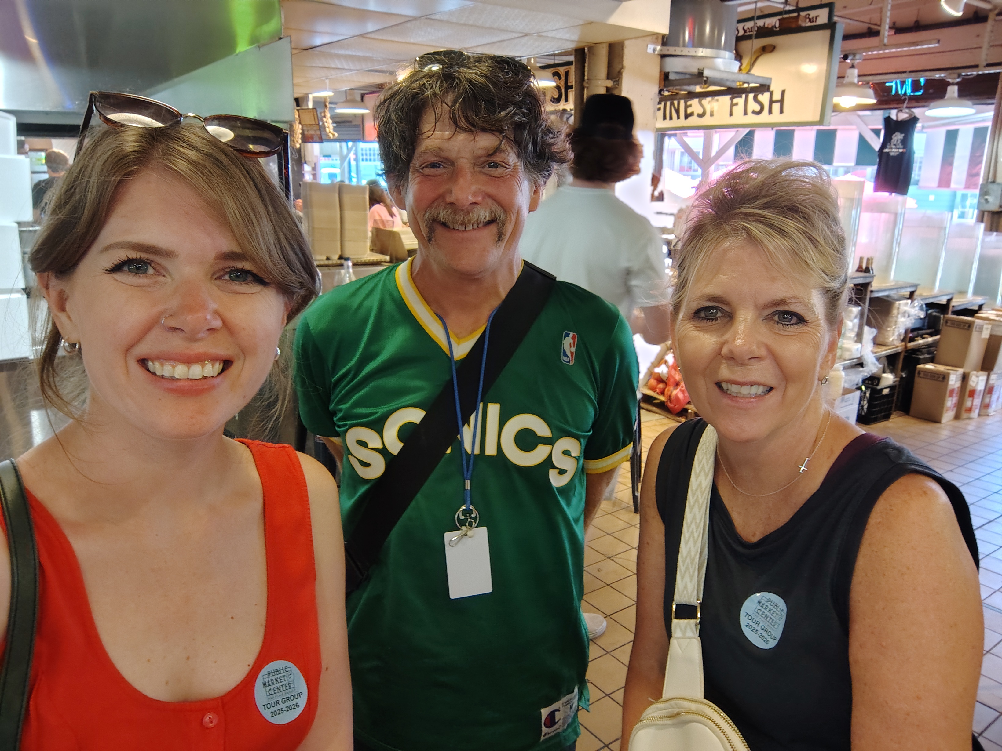 Two white women pose with a white man. One woman has brown hair and a red dress, the other has blond hair and a dark grey top. The man wears a green 